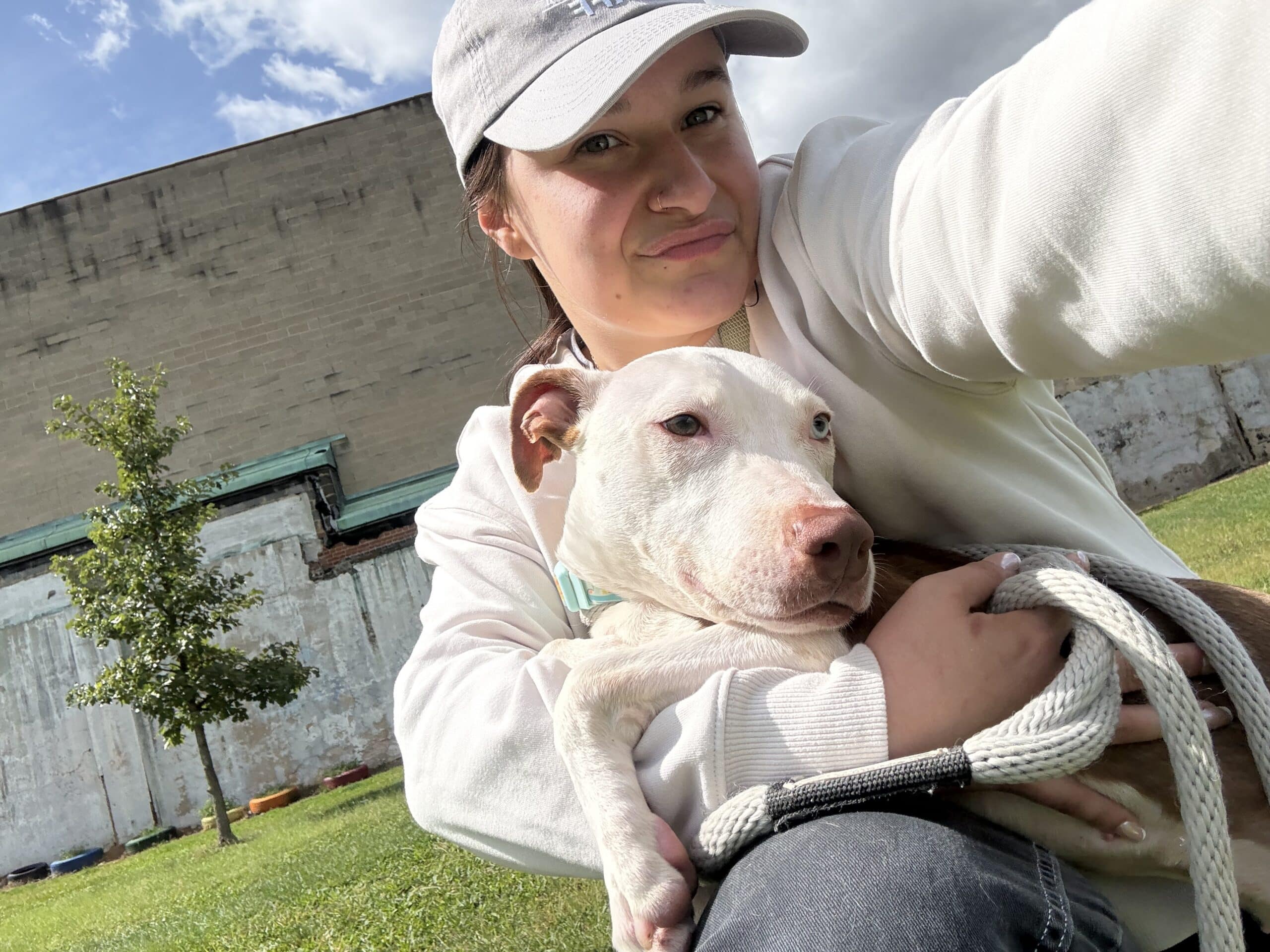 Young woman taking a selfie with a white pit bull outdoors in front of a shelter building
