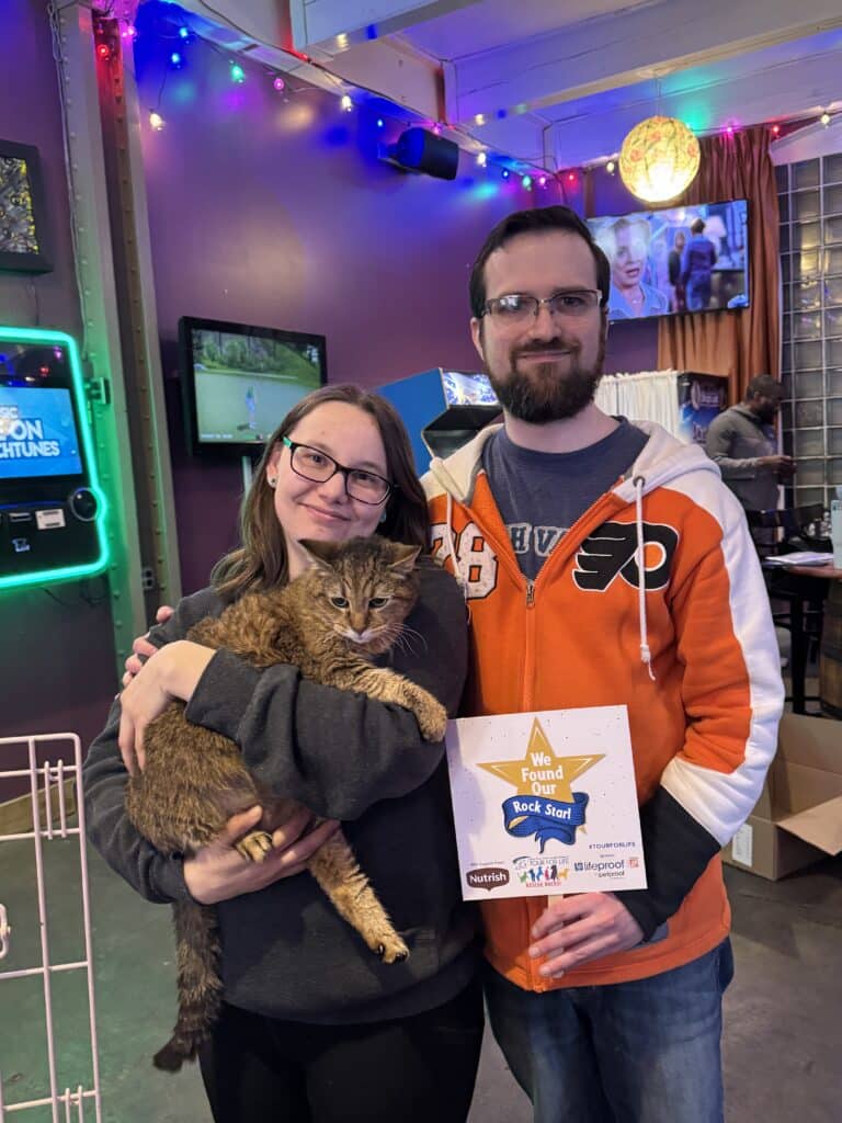 Young couple — man in Flyers jersey — holding a brown tabby cat and 'We Found a SPCA Best Friend' star sign in their liv
