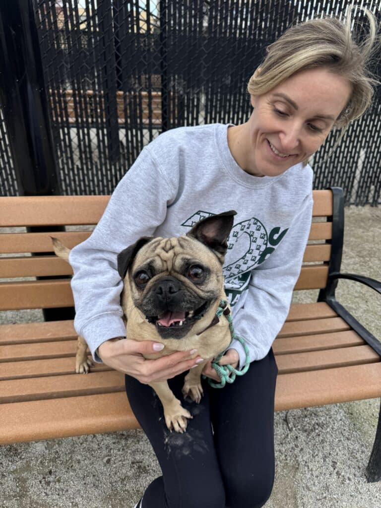 Mujer rubia sonriente con una sudadera gris, con un pug de color beige en el regazo, sentada en un banco frente al refugio