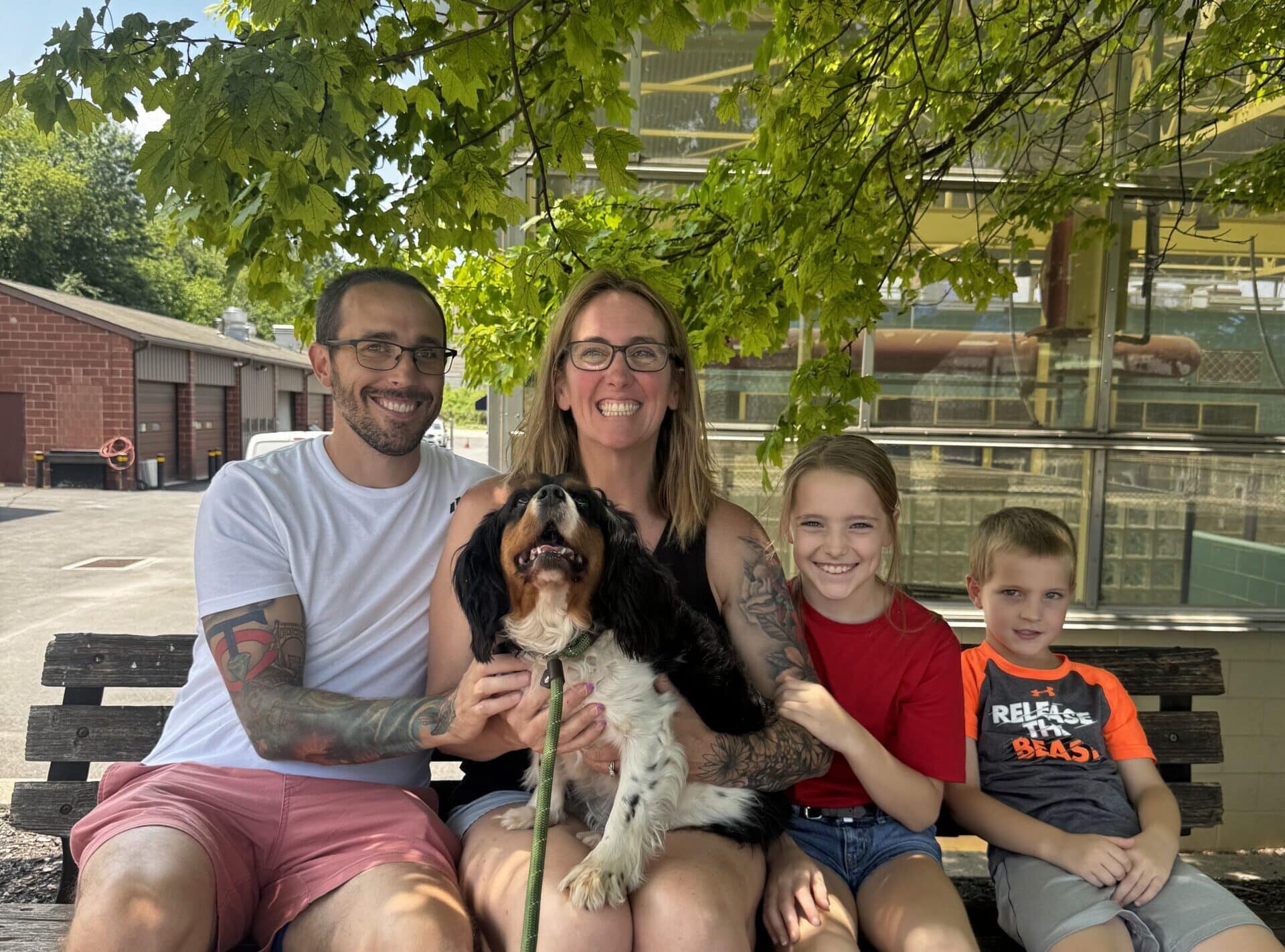 Young family of four with two kids sitting under a leafy tree holding a small scruffy terrier mix — beautiful adoption d