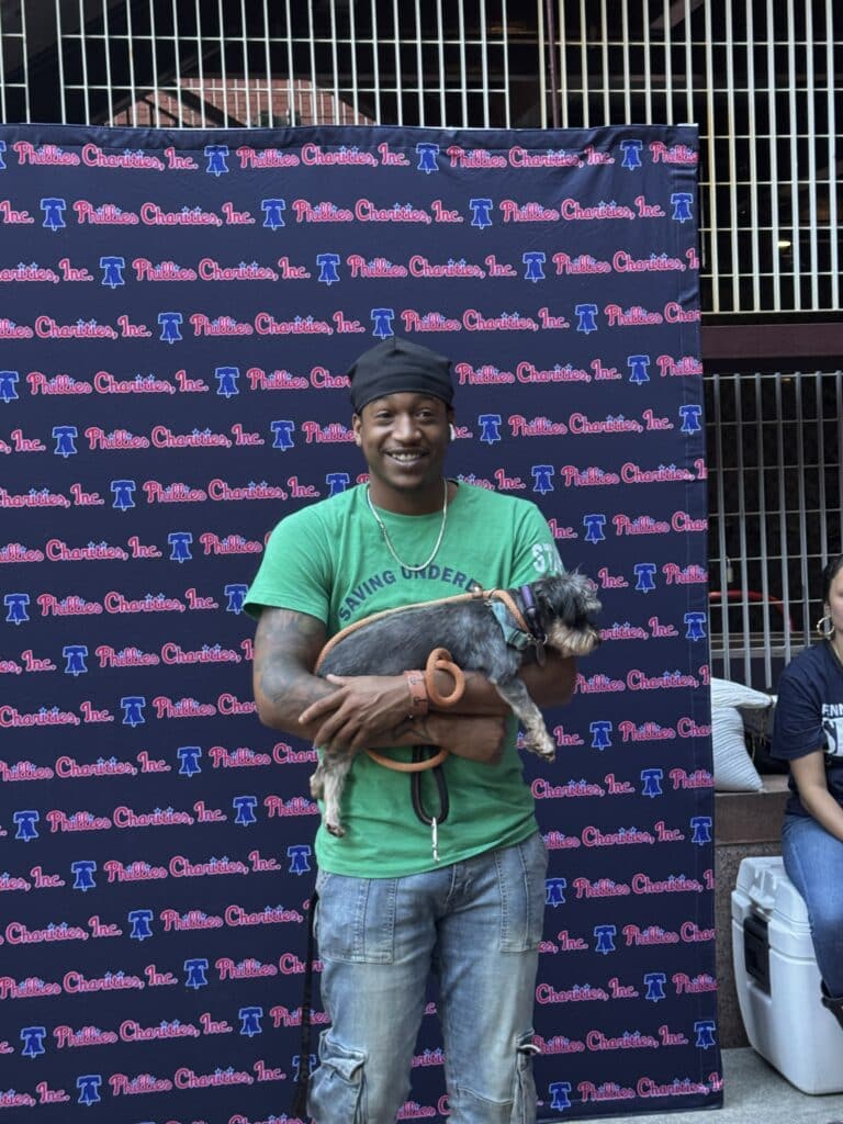 Smiling man in green PSPCA 'Saving Modern' t-shirt holding a small grey Chihuahua mix at the Phillies stadium photo back