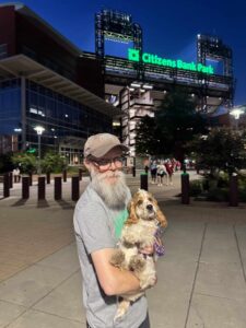 Woman in ball cap holding a small scruffy terrier mix outside Citizens Bank Park at dusk, stadium sign glowing green beh