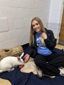 Una joven con una camiseta azul de la PSPCA sentada con un conejo blanco sobre un lecho de heno en el refugio