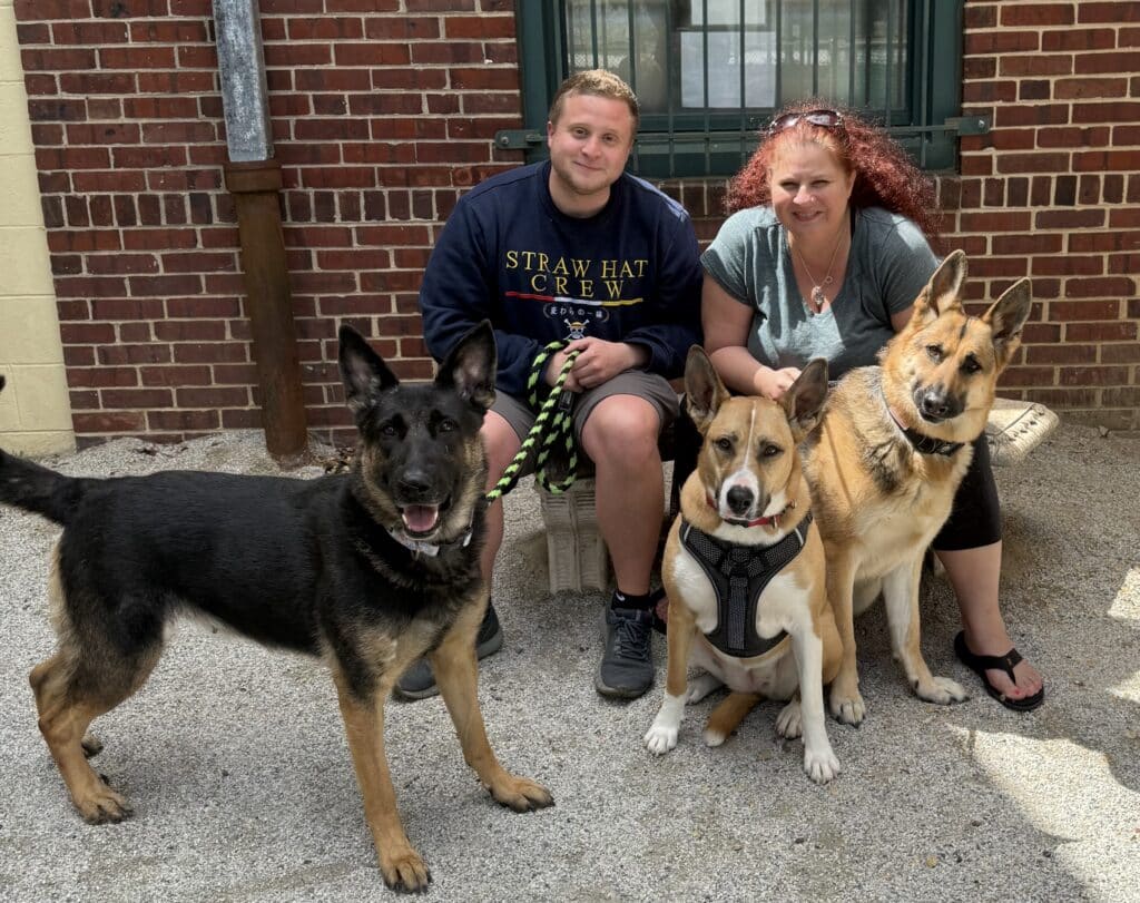 Young couple sitting outside with three German Shepherds — happy multi-dog adoption family photo by brick building